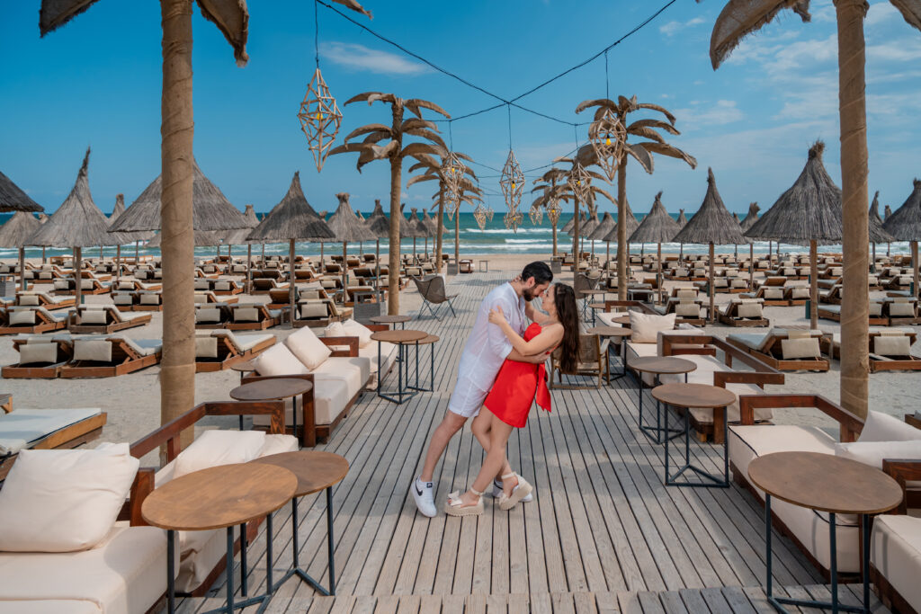 Cuplu într-o ședință foto pre-wedding pe plajă, într-un beach club elegant, cu șezlonguri și palmieri